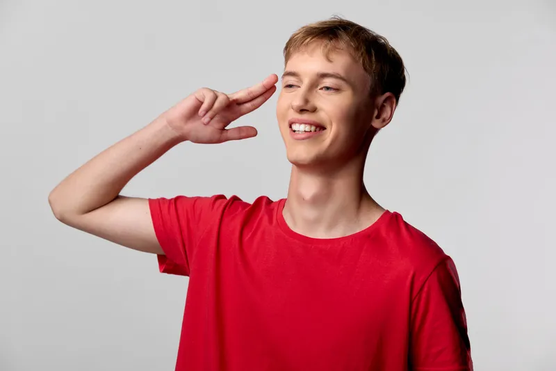 Smiling young man in a red t shirt saluting with fingers near temple, cheerful casual pose conveying greeting, confidence and friendly energy for lifestyle, fashion and stock photography, suitable