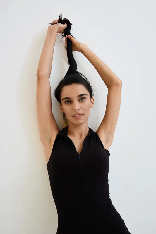 Young woman with long black hair in black sleeveless outfit stretching arms upward against white background, showcasing natural beauty and casual style.