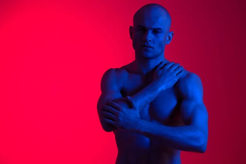 Portrait of a man posing with arms folded, surrounded by contrasting blue and red lights, creating a striking black and white aesthetic.