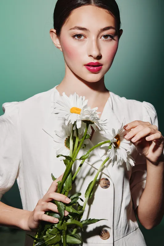 Young woman with makeup modeling holding white daisies. She wears elegant white clothing with decorative buttons on green background. Beauty concept with floral and fashion details.
