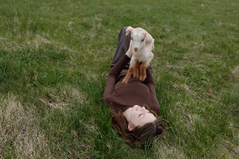 Young woman lying on green grass holding a baby goat, enjoying a peaceful outdoor moment, cozy sweater, candid lifestyle scene, sunny natural light, gentle animal companionship on a meadow.