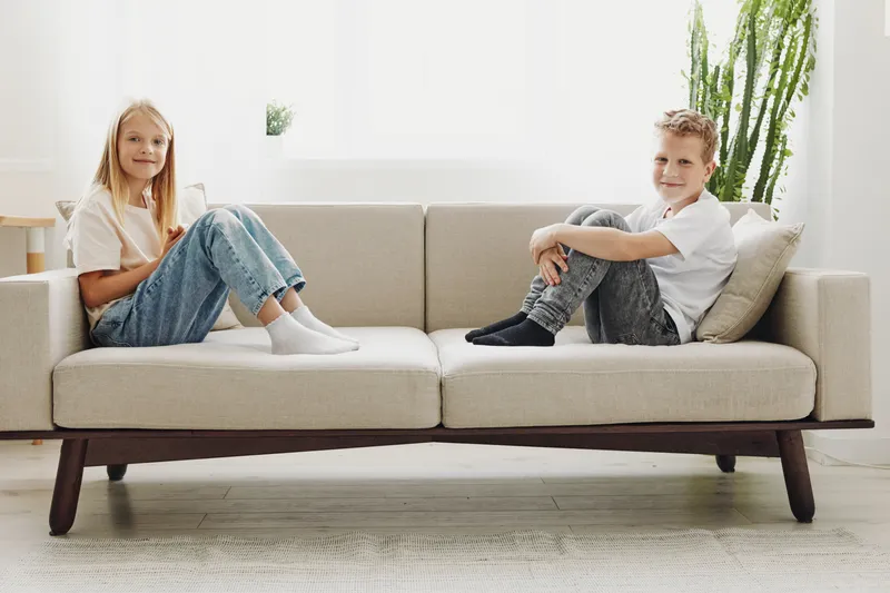 Two children sitting on opposite ends of a modern beige sofa in a bright room with natural light and minimal decor. Concept of family, childhood, and home interior.