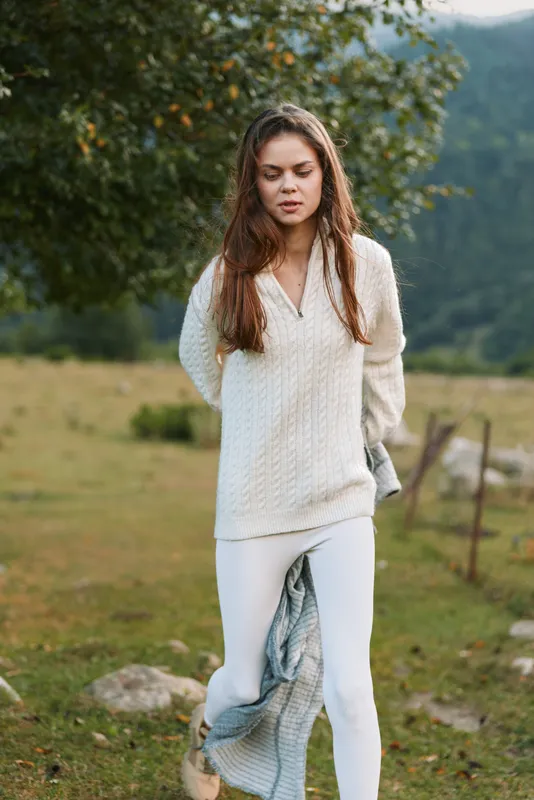 Young woman in white sweater and pants walking outdoors in nature with a blanket in hand. Relaxed lifestyle scene with green trees and mountains in background.