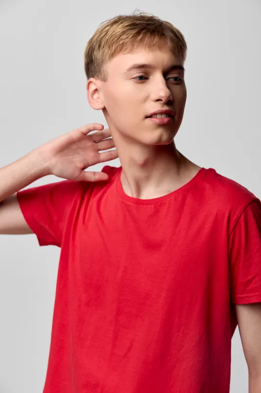 Man in a red shirt strikes a casual studio pose, head turned to the side, highlighting relaxed fashion, confident attitude and contemporary style, with a clean background and soft lighting for