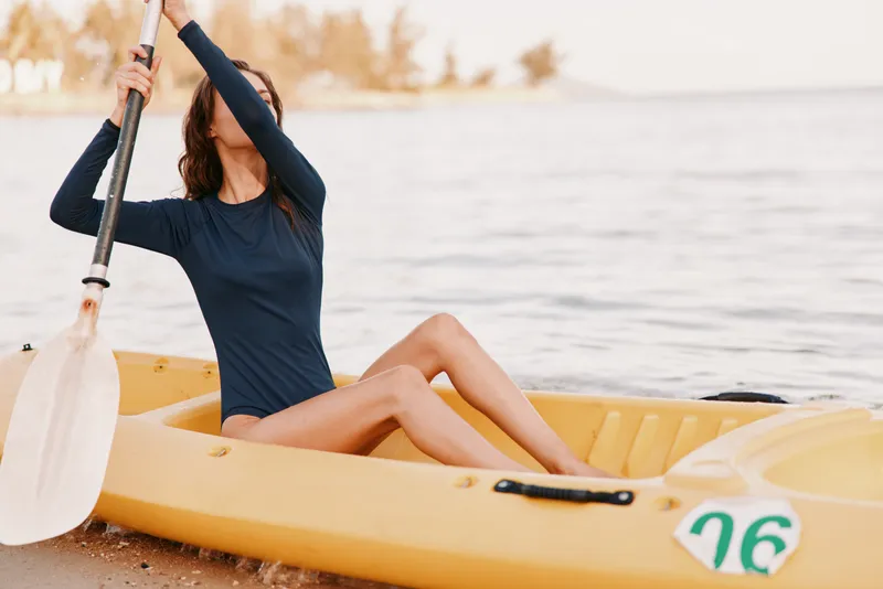 A woman paddles a bright yellow kayak across calm water, raising an oar overhead as the sun casts warm light on the scene. Her navy long sleeve swimsuit contrasts with the vivid craft.