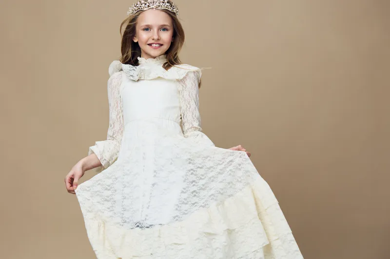 A joyful young girl in a light lace dress and crown, smiling and elegantly posing on a neutral beige background, perfect for celebrating childhood and innocence
