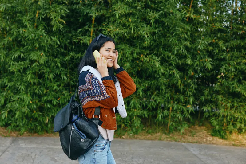 Phone conversation with a cheerful woman outdoors, backpack slung over a casual jacket and denim, standing by a green hedge on a sunny day, street style moment