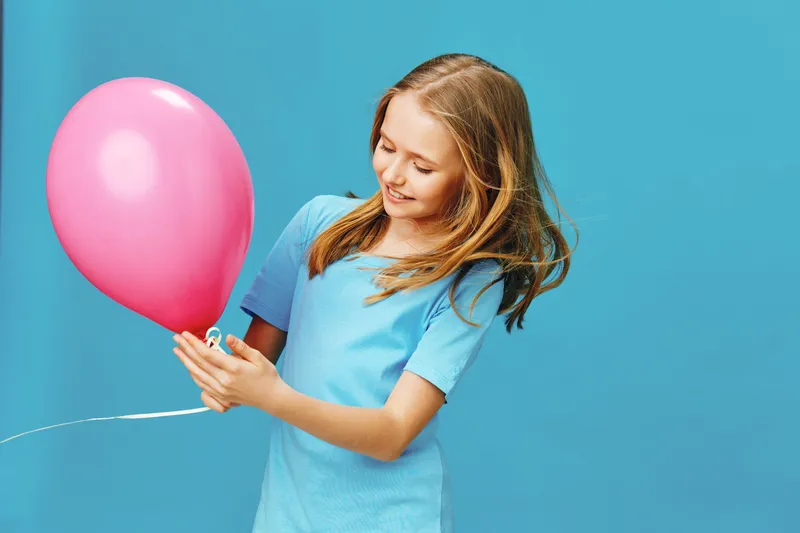 Happy young girl with a pink balloon against a vibrant blue background, expressing joy and innocence in a playful, cheerful moment