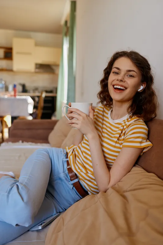 A smiling woman relaxes at home, seated on a sofa with a mug in a striped shirt. Cozy living room scene conveys casual comfort, warmth, and modern woman lifestyle.