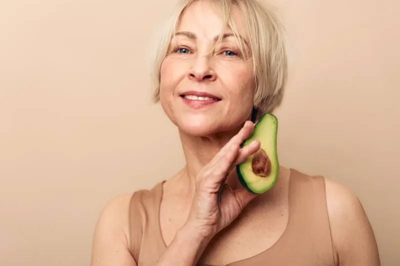 Smiling mature woman holds fresh avocado near face on beige background. Concept of healthy lifestyle, natural beauty, nutrition, wellness, and skin care for older adults.