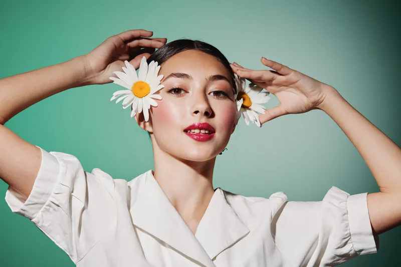 Young woman with moist dewy skin wears white blouse and places large daisies in her hair in creative fashion beauty concept on green background studio setting.