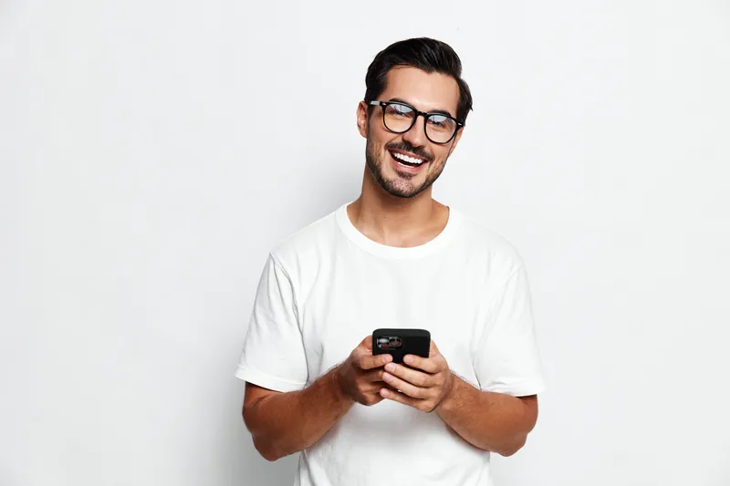 Smiling man wearing glasses holds smartphone in hands, looking at camera with happy expression, standing against plain white background. Lifestyle concept