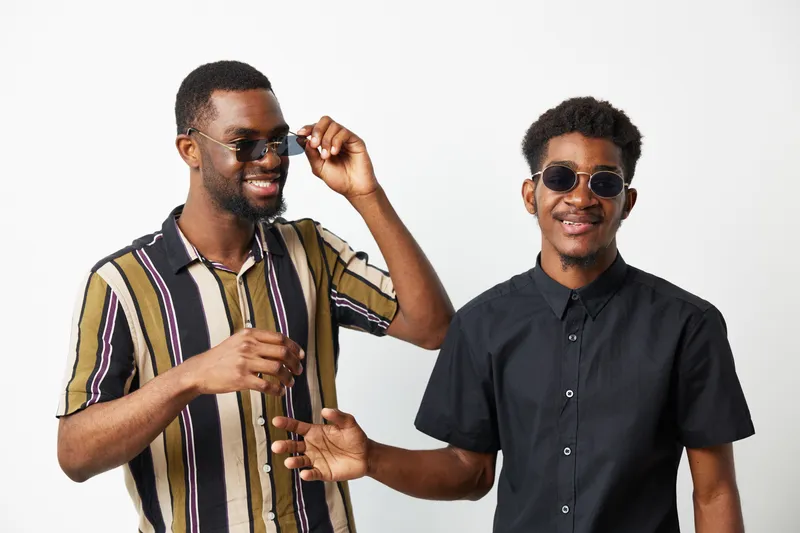 Two smiling black men wearing sunglasses, showcasing a fun and relaxed vibe. They are dressed in stylish shirts, embodying summer's joyful spirit.