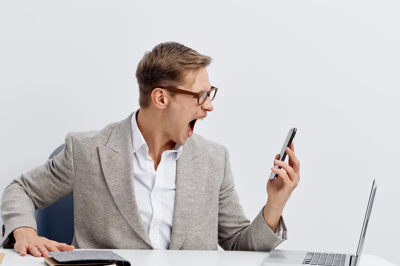 Confident man using smartphone in office, wearing glasses and formal blazer, expressing excitement or surprise while sitting at desk with laptop, white background, professional business environment, t