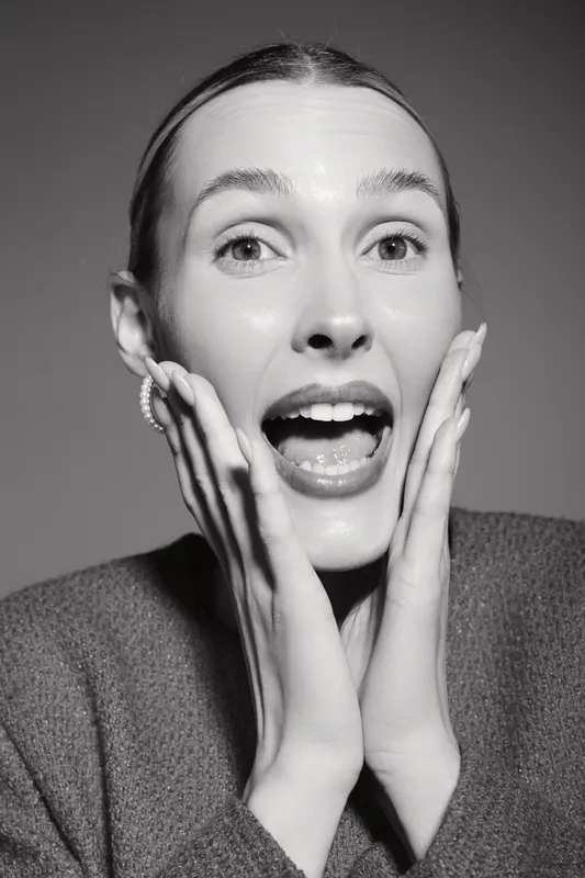 Surprised Caucasian woman with fair skin and slicked hair holds face with hands, open mouth and wide eyes. Concept of astonishment, excitement, and emotion in studio.