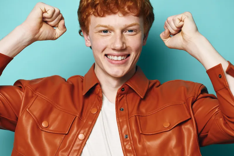 Young man with red hair and freckles smiling and raising fists in excitement wearing a brown leather jacket and white shirt against a blue background portrait.