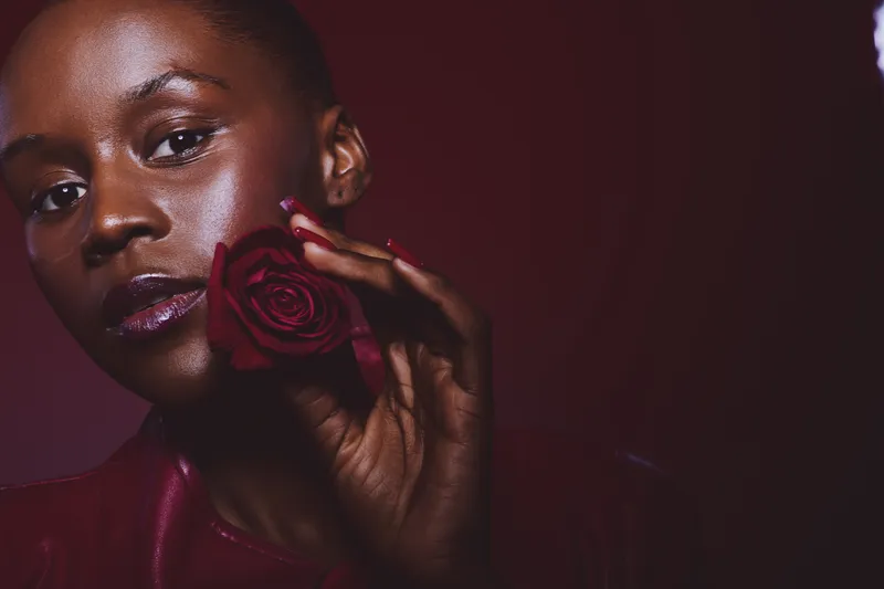 Close-up portrait of a dark skinned African woman holding a red rose near her face, wearing dark red clothing. Concept of beauty, elegance, and natural skincare in studio lighting.