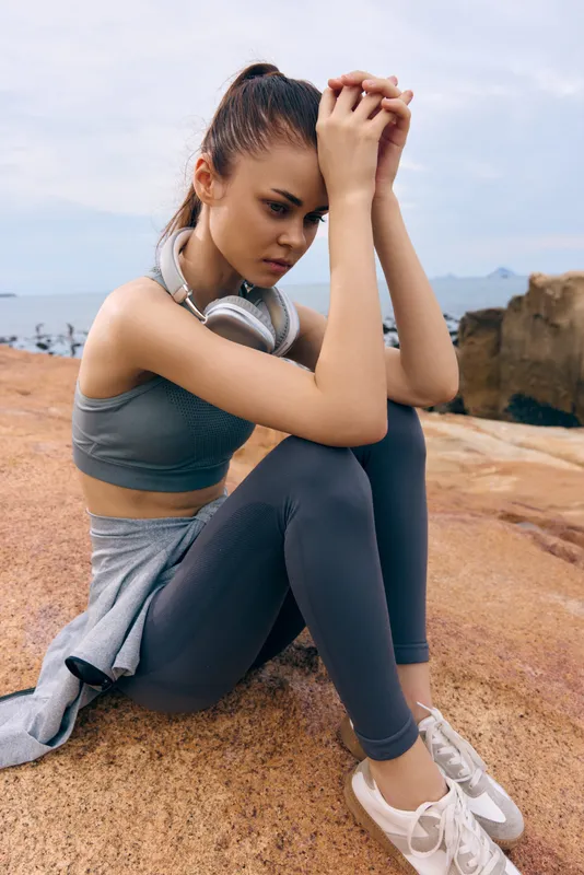 Thoughtful young woman in sporty attire sits on rocky surface, reflecting on fitness journey amidst a serene coastal background, wearing headphones