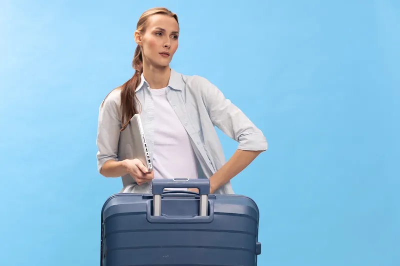 Young woman with a suitcase and laptop stands against a blue background. She looks thoughtful while preparing for travel or business trip, concept of modern travel and technology use.