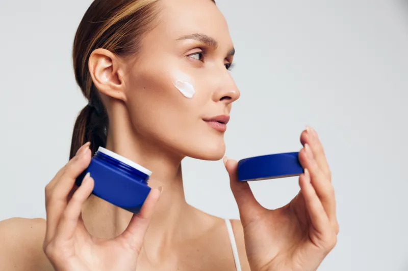 Young woman with brunette hair applies face cream from a blue jar on her cheek while looking sideways against a plain light background with a calm expression.