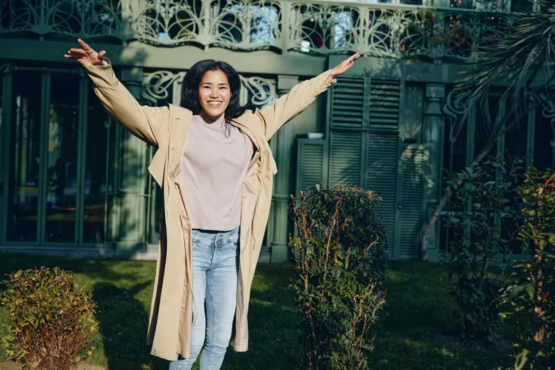 Joyful woman with arms wide open in a light trench coat outdoors, greenery and decorative iron gate behind, casual fashion moment captured in daylight