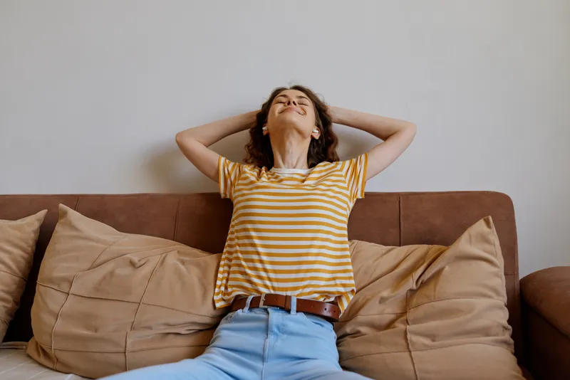 Relaxed woman lounging on a sofa at home, wearing a striped shirt. Casual lifestyle scene emphasizing comfort, serenity, and wellbeing in a cozy living space. Warm lighting enhances mood.