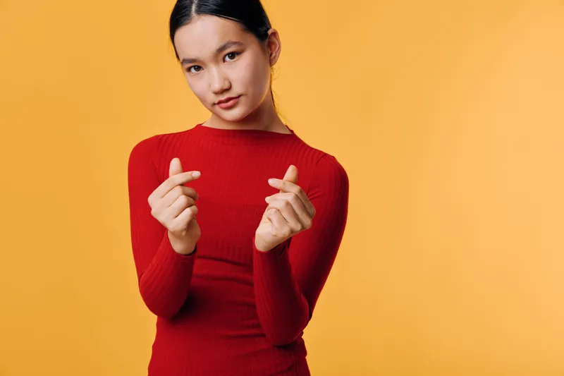 Young woman in red sweater making finger heart gesture with hands, posing confidently against orange background. Asian girl, casual style, expression and sign for love or affection.