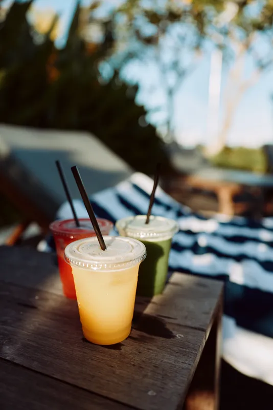 Three colorful beverages on a wooden table outdoors, sunlight and shadows creating a relaxing summer vibe. Plastic cups with straws sit near a striped towel, suggesting a casual backyard gathering