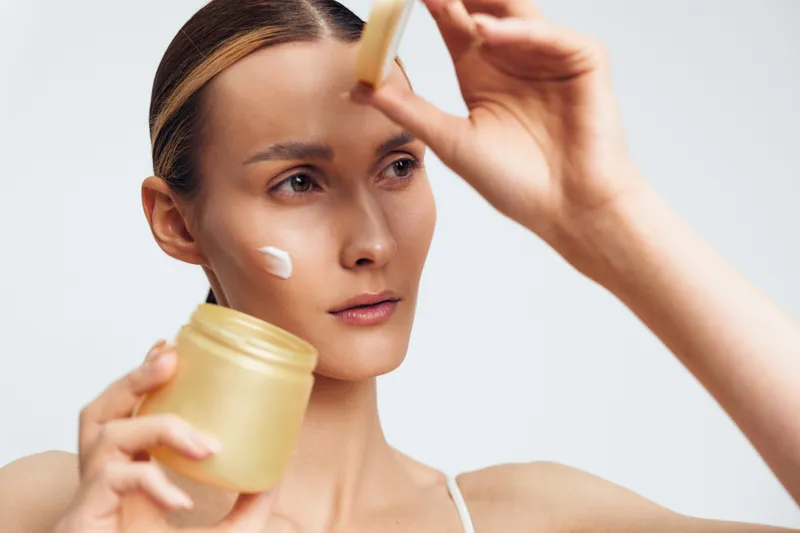 Young woman applies cream on her face with a brush holds jar of skincare product has clean fresh skin and focused expression isolated on white background.