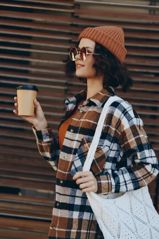 Young woman holding coffee in paper cup, wearing beanie, sunglasses and plaid shirt with tote bag, smiling in urban street scene; authenticity, candid lifestyle, golden hour glow and emotional