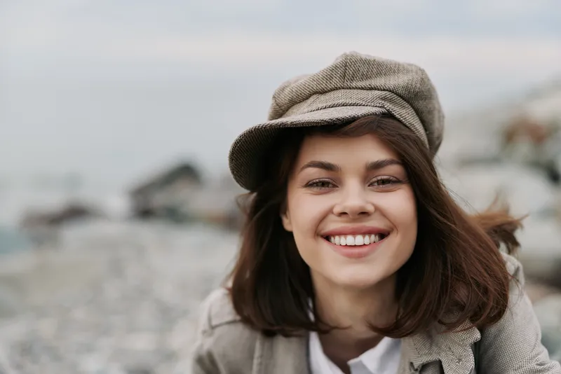 Smiling young woman wearing a brown hat and coat stands outdoors on a rocky beach. Natural light highlights her happy expression and casual style. Concept of joy and nature.