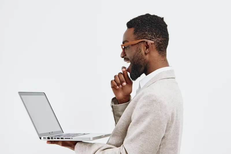 Young smiling African American man wearing glasses and beige sweater using a laptop computer in profile view isolated on white background. People lifestyle concept.