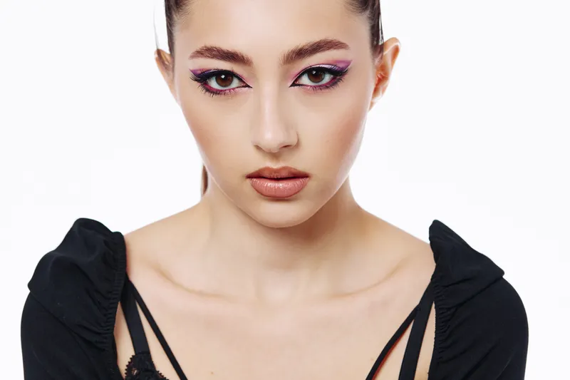 Young woman with vivid purple eyeliner and flawless skin poses confidently in a modern black top. Studio close-up with white background. Beauty fashion concept featuring bold makeup.