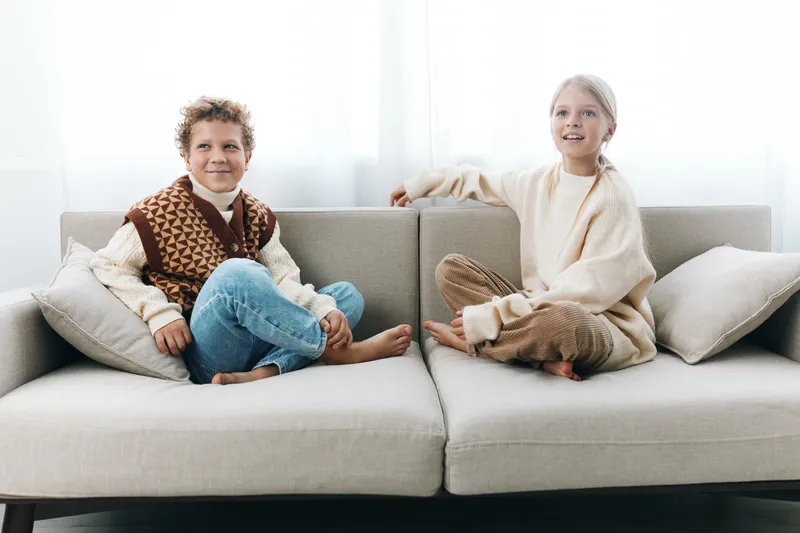 Two children sit on a light sofa wearing cozy sweaters, smiling softly as natural light fills a bright living room and creates a warm, inviting scene.
