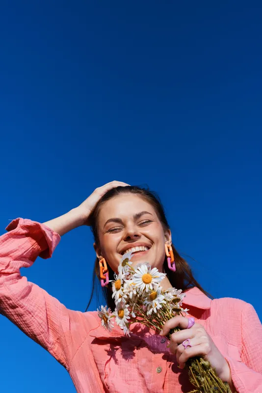 Young woman in pink shirt joyfully holding daisies against a clear blue sky, radiating happiness and a fresh, vibrant vibe