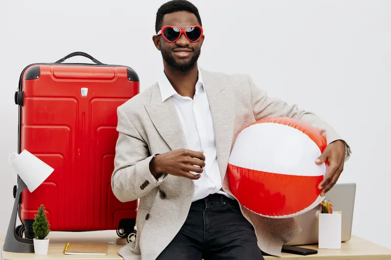 Man holding beach ball in indoor studio with travel luggage, wearing sunglasses, cheerful expression, casual summer style. Bright, vibrant scene emphasizing vacation and leisure atmosphere.