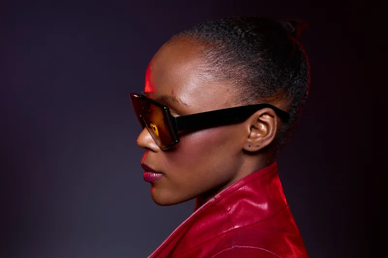 Profile of a confident black woman wearing large sunglasses and a red leather jacket against a dark background. Concept of fashion, style, confidence, and urban elegance.