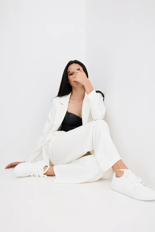 Young woman sitting on floor against white wall in stylish white suit and black top, casual pose with hand near face, minimalistic studio background, modern fashion.