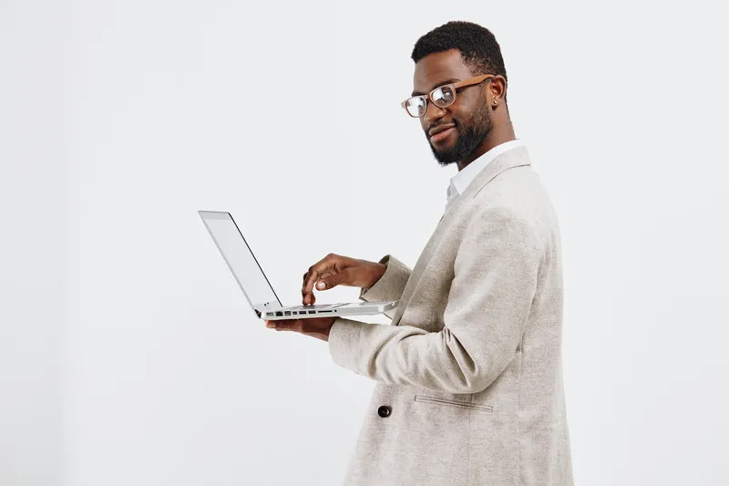 Confident young African American man in beige blazer holding laptop with smiling expression, looking away, isolated on white background, studio portrait. Business lifestyle concept, professionalism, s