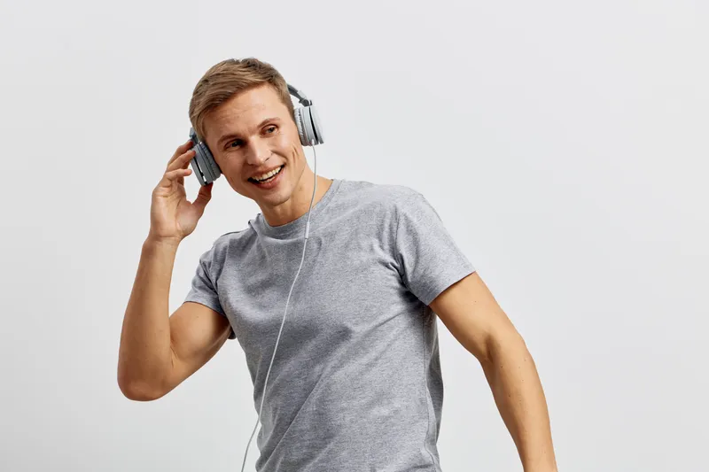 Smiling young man wearing casual gray t-shirt listening to music with headphones, looking happy, cheerful, engaging in leisure activity, isolated on plain light background, studio portrait. People lif