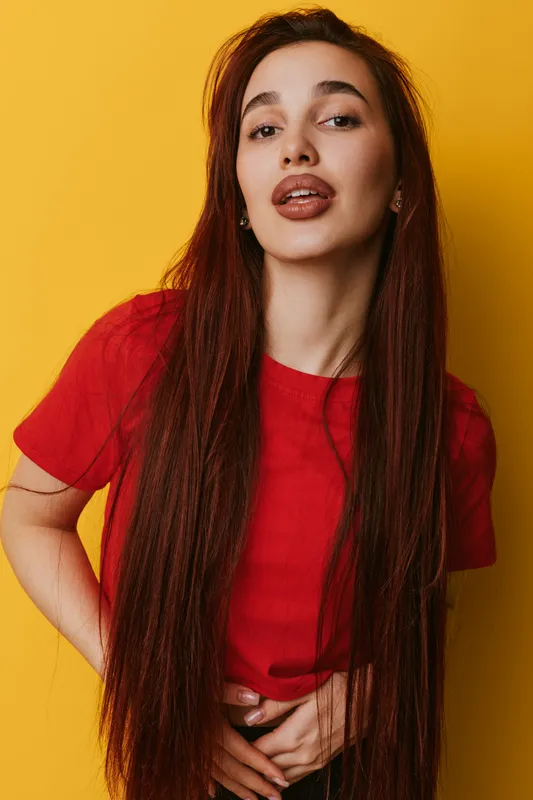 Young woman with long brown hair wearing red t shirt posing with hands on waist against yellow background. Female model looking confident and stylish with natural makeup and earrings.