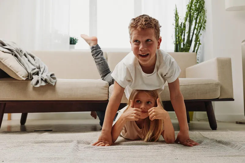 Two playful children in casual clothes have fun on a carpet at home, boy balancing on girl who shows thumbs up. Natural light, cozy living room, concept of sibling joy and family bonding.