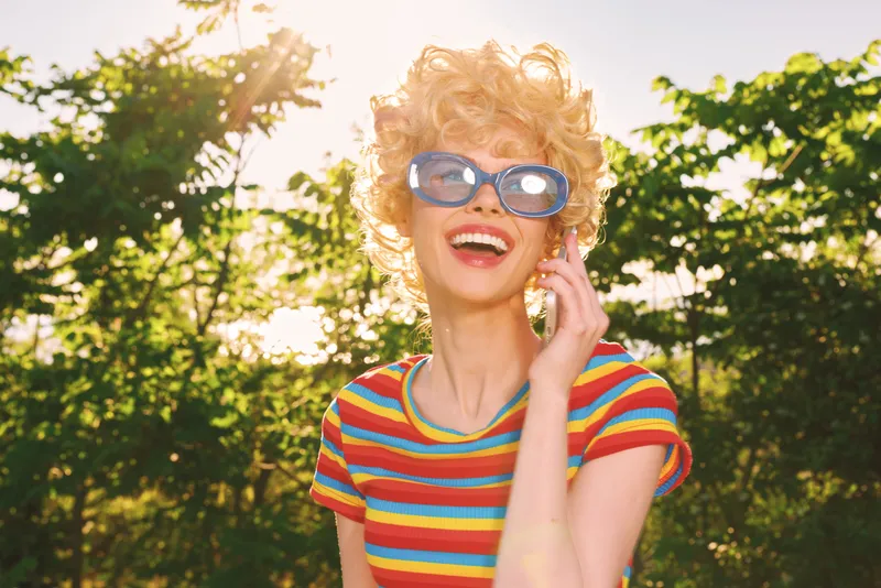 Young woman with curly blond hair laughs outdoors in a colorful striped shirt, enjoying a sunny moment with retro film color and carefree energy.