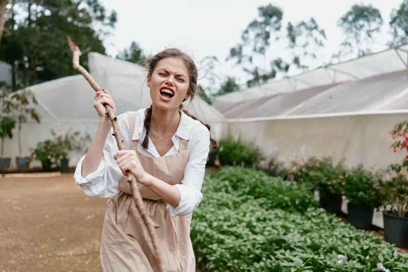 Young woman in an apron passionately gardening, holding a stick, showcasing determination in a vibrant greenhouse filled with lush greenery