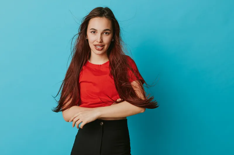 Young woman with long hair wearing red shirt and black pants posing with crossed arms smiling on blue background. Casual style, confident female model with natural makeup and relaxed posture.