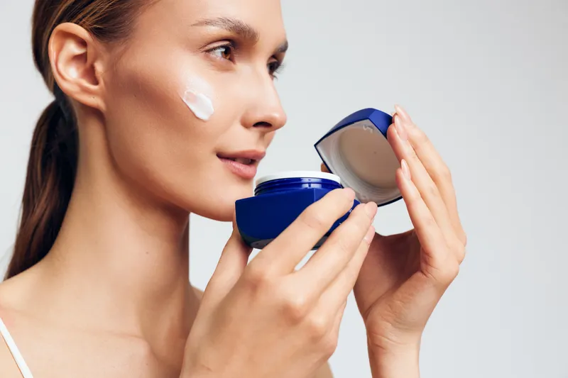 Young woman applies moisturizing cream on face holds open jar with white cosmetic product looks focused on skincare routine isolated on light background.