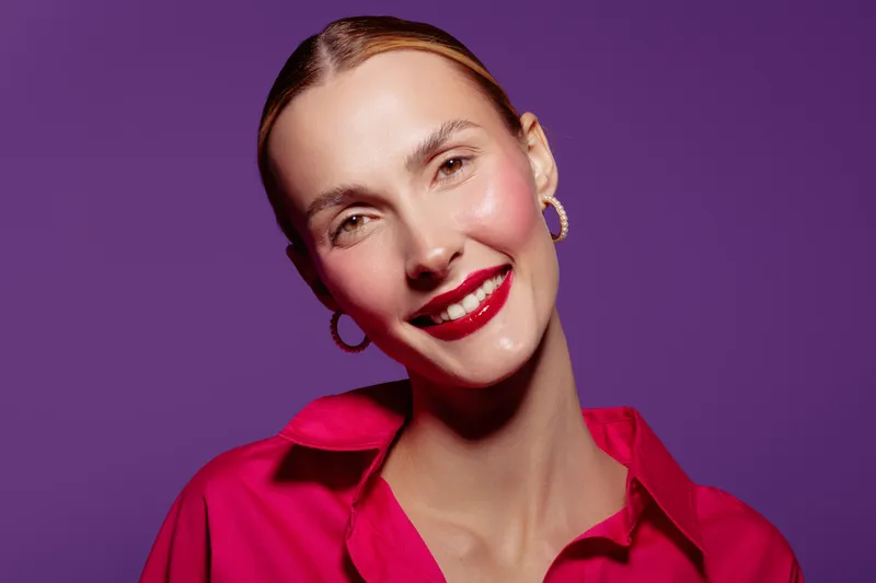 Smiling woman with red lipstick and gold hoop earrings poses against purple background. She wears a bright red shirt, showcasing beauty, fashion, and confident expression.