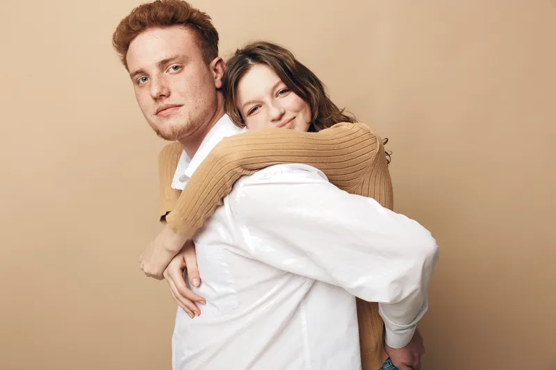 Young couple in casual clothes share a warm embrace against beige background. The woman smiles happily while hugging the man from behind. Concept of love, affection, and togetherness.