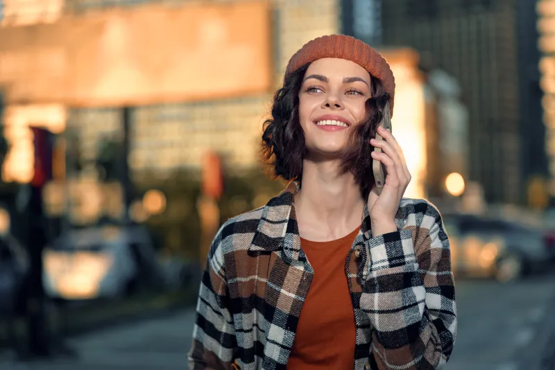 Woman on phone smiling in urban street during golden hour glow, candid lifestyle portrait with authenticity and mindful living vibe, emotional storytelling through natural expression.