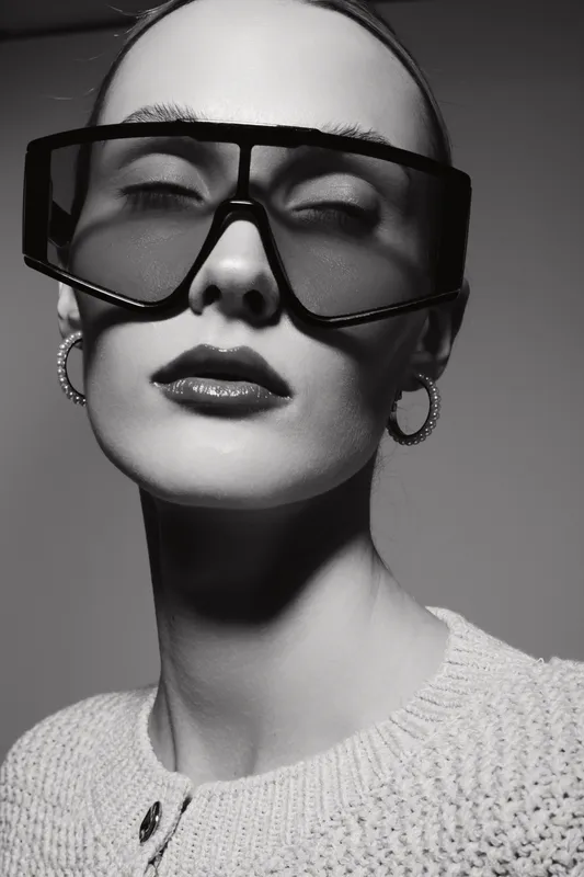 Elegant white woman with pale skin poses wearing large sunglasses and hoop earrings in black and white studio portrait with stylish sweater and smooth makeup.